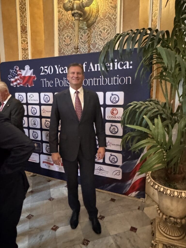 Peter Wachowski posing in front of the "250 Years of America: The Polish Contribution" sponsor recognition display at the Polish American Congress event.